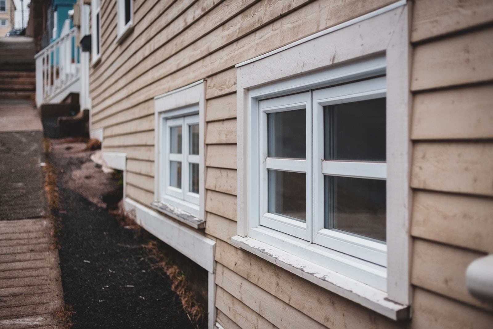 Two white basement windows set low in tan siding along a walkway