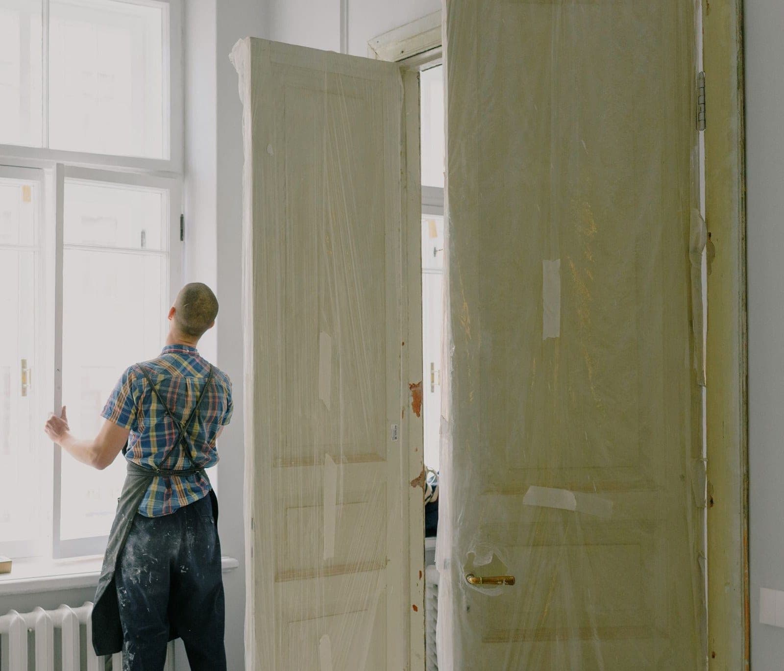 Installer in an apron fitting a tall window inside a bright room, drill on the sill